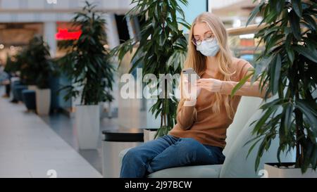 Caucasian busy woman in medical mask sitting on sofa business chatting. Masked girl female ill shopping mall center sitting on couch looking at mobile Stock Photo