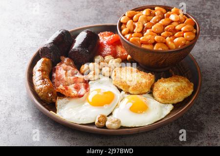 Full fry up English breakfast with fried eggs, sausages, bacon, black pudding, beans, tomato closeup in the plate on the table. Horizontal Stock Photo