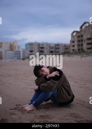 non binary young woman alone on the beach sitting on the sand Stock Photo