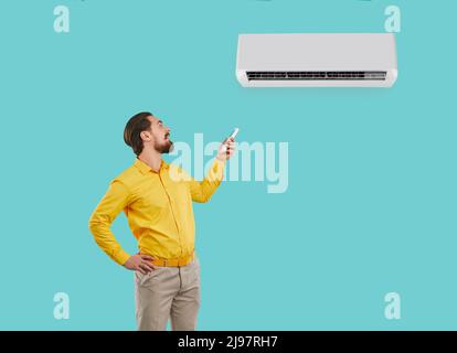 Happy man holding remote control controls or adjusts newly installed wall-mounted air conditioner. Stock Photo