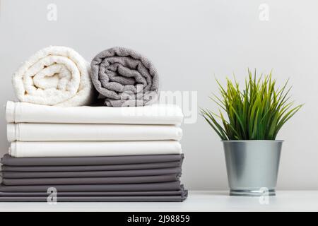 A stack of gray and white linens, bed sheets and a folded roll of towels a plant in a pot on a table. Stock Photo