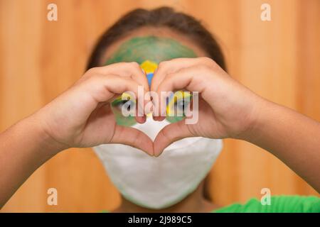 child with mask with his face painted with the flag of brazil, making a heart with his hands. Stock Photo