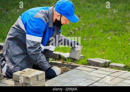 Bricklayer in work clothes sits on sidewalk and lays out paving slabs. Sight of working man in open air. Professional builder makes arrangement of territory summer day. Real scene. Stock Photo