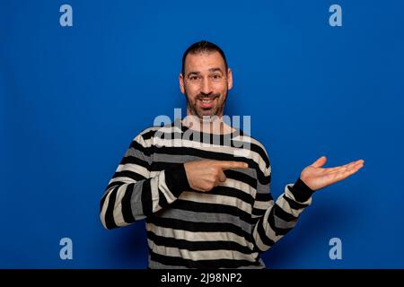 Surprised man in striped sweater pointing left with his finger, shouting WOW, isolated on blue background with copy space for your text. shock content Stock Photo