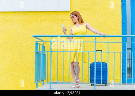 Beautiful red-haired woman stands with a blue suitcase and uses a smartphone on a yellow background. Stock Photo