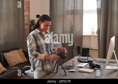 Stylish young woman with afro braids wearing casual outfit standing at desk checking and adjusting camera settings to shoot video for blog Stock Photo