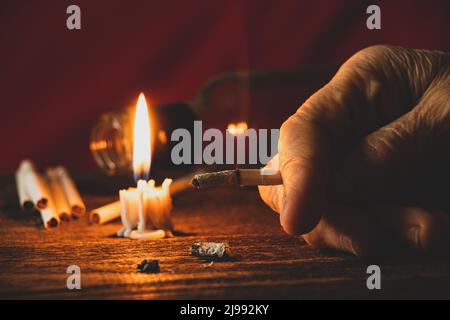 old woman holds a cigarette by candlelight in the dark at home at the  table, smokes at home in the dark, nicotine and a bad habit, cigarettes and  canc Stock Photo -