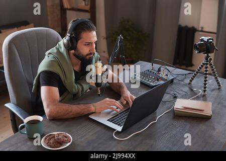 Horizontal high angle portrait of modern Middle Eastern male influencer sitting at desk in loft room working on laptop Stock Photo