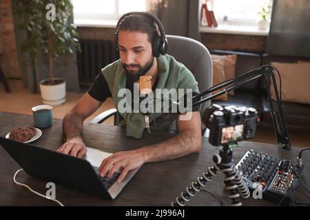 Horizontal high angle shot of handsome bearded young Middle Eastern man wearing casual clothes sitting alone in loft room creating content for vlog Stock Photo