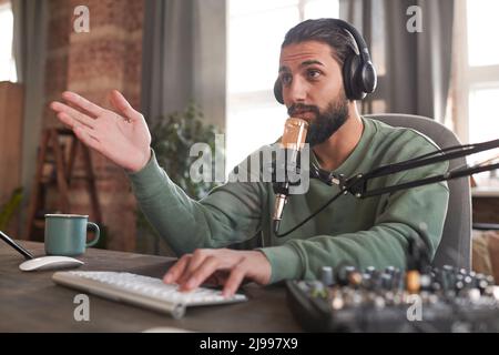 Horizontal medium portrait of handsome young Middle Eastern man wearing headphones sitting at desk speaking to followers during stream Stock Photo