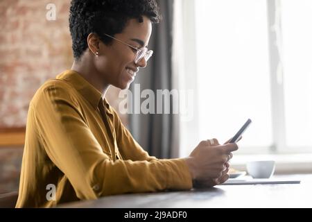 Smiling African woman in eyeglasses sit at table using smartphone Stock Photo