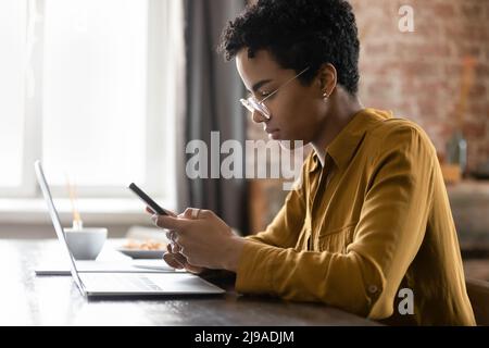 Serious African woman sit at desk with laptop using smartphone Stock Photo