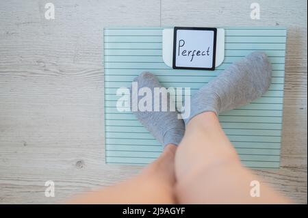 Faceless woman measures weight on the floor scales. Top view of women's feet in gray socks on the scales with the word perfect. The inscription on the Stock Photo