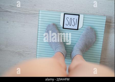 The fat woman is weighed. A top view of female feet in gray socks stands on an electronic scale. SOS inscription on the display of the floor scale. Stock Photo