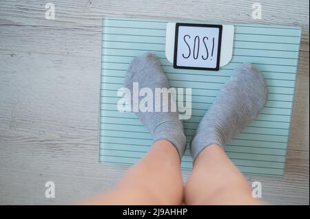 The fat woman is weighed. A top view of female feet in gray socks stands on an electronic scale. SOS inscription on the display of the floor scale. Stock Photo