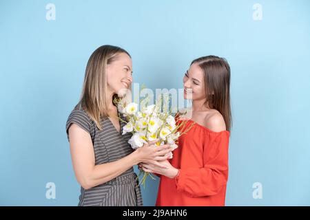 Portrait charming pretty attractive mother and daughter with big bouquet of aromatic flowers happy smiling in red and gray dresses, isolated on blue background. Family and togetherness concept Stock Photo