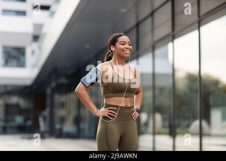 Young black woman in sports outfit listening to music in earphones, using mobile device during jogging on city street Stock Photo