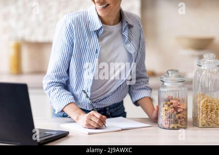 Young housewife watching video on laptop and writing new recipe, standing in kitchen interior, crop Stock Photo