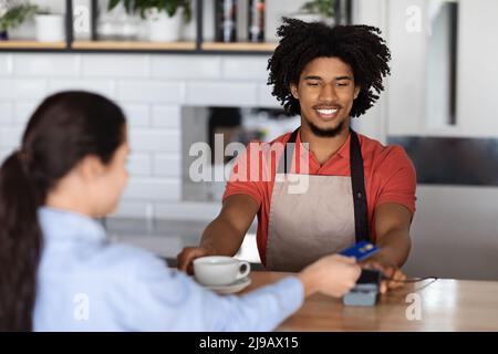 Smiling curly young black man bartender in apron at bar counter gives terminal to lady client with credit card Stock Photo