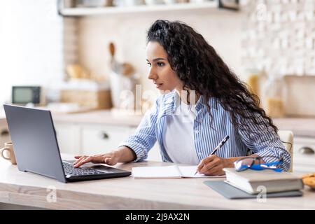 Concentrated woman in casual working on laptop in minimalist kitchen interior, looking at screen and taking notes Stock Photo