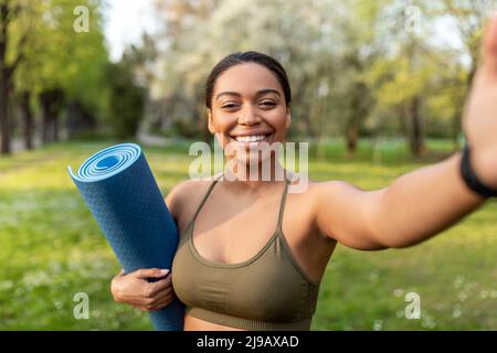 Millennial black woman in sportswear holding yoga mat, taking selfie on smartphone after outdoor training Stock Photo