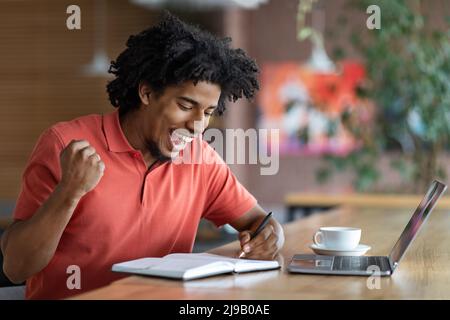 Glad happy excited curly handsome young black man make notes in notebook, victory gesture in cafe interior Stock Photo
