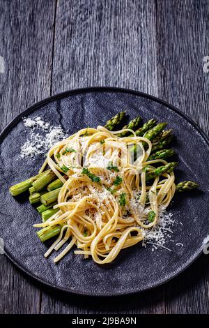 roasted asparagus spears with spaghetti sprinkled with shredded pecorino cheese and parsley on black plate on dark wood table, vertical view from abov Stock Photo