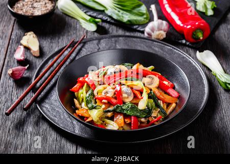 Stir Fry Vegetables of bok choy, carrots and chinese stir fry sauce in black bowl on black wooden table with ingredients on background, mushrooms, oni Stock Photo