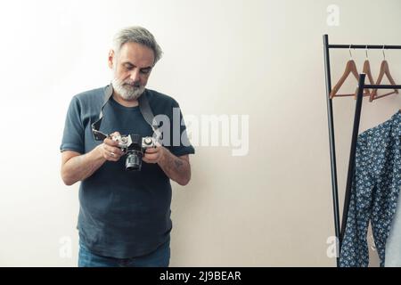 Medium studio shot. Elderly casually dressed grey-haired man holding a professional camera in his hands, standing near a clothes rack. High quality photo Stock Photo