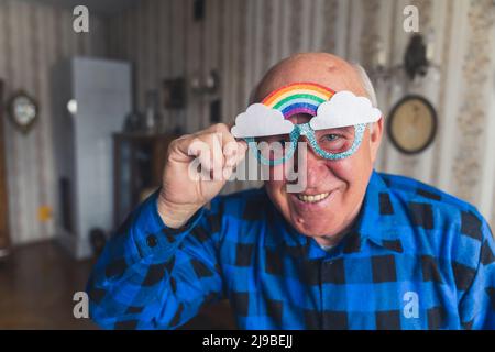 medium closeup portrait of a joyful senior Caucasian man with rainbow-shaped funny glasses at home senior people support concept. High quality photo Stock Photo
