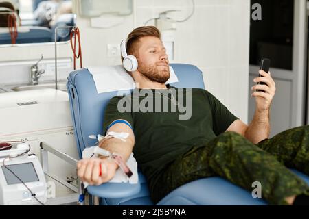 Portrait of smiling military man giving blood while laying in chair at blood donation center and holding smartphone, copy space Stock Photo