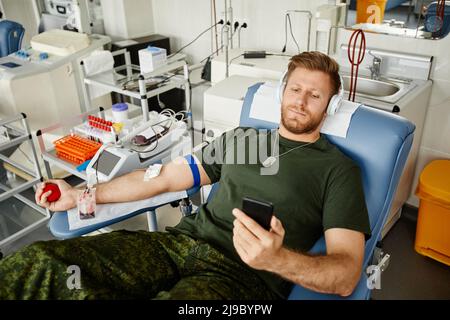 High angle portrait of military man giving blood while laying in chair at blood donation center and watching videos at smartphone, copy space Stock Photo