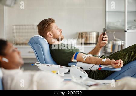 Side view portrait of military man giving blood while laying in chair at blood donation center and watching videos at smartphone Stock Photo