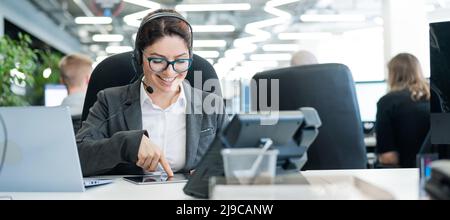 Beautiful caucasian woman in headset is holding a digital tablet while standing in open space office. A friendly female support service operator Stock Photo