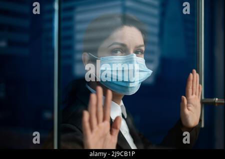 A sad quarantined woman in a business suit leaned against the window. Caucasian girl in a mask on isolation put her hands on a glass door. Maintaining Stock Photo