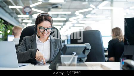 Beautiful caucasian woman in headset is holding a digital tablet while standing in open space office. A friendly female support service operator Stock Photo