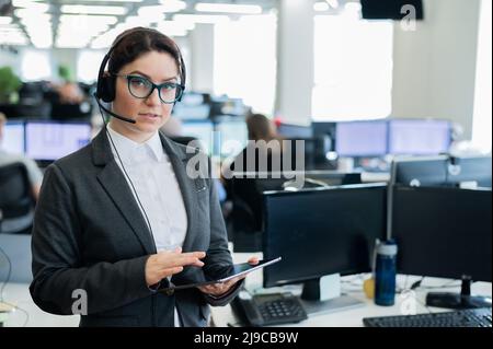 Beautiful caucasian woman in headset is holding a digital tablet while standing in open space office. A friendly female support service operator Stock Photo