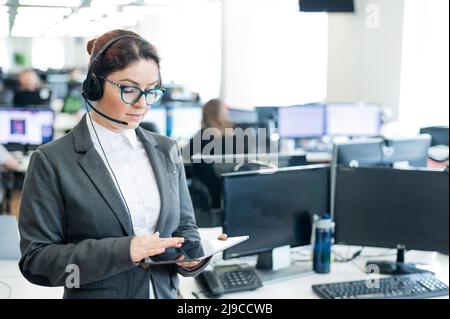 Beautiful caucasian woman in headset is holding a digital tablet while standing in open space office. A friendly female support service operator Stock Photo
