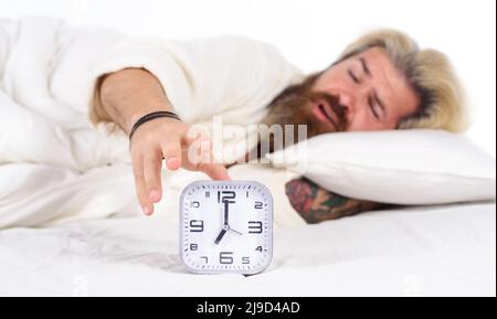 Morning awakening. Sleepy bearded man turning off alarm clock. Male in bed. Early wake up, insomnia. Stock Photo