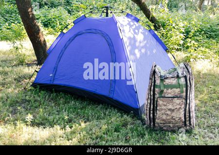 Defocus blue open tourist tent standing on green nature background. Army backpack. Tourism concept. Summer vacation in forest, camping. Blurred. Out o Stock Photo