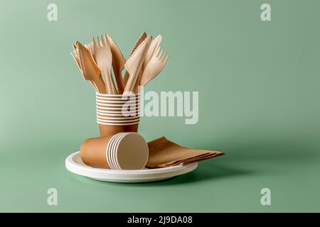Stack of eco-friendly disposable tableware. Wooden forks and knives, paper cups and plates against green background. Biodegradable cutlery and dishes. Stock Photo