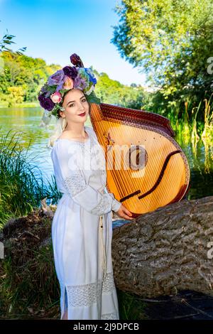 Young Ukrainian woman in national dress plays a bandura by the river on a summer sunny day. Stock Photo