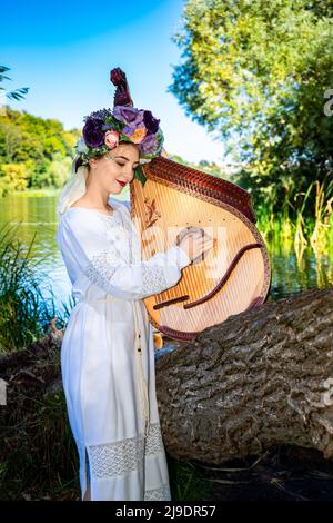 Young Ukrainian woman in national dress plays a bandura by the river on a summer sunny day. Stock Photo