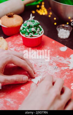 Christmas tree shaped cupcakes with snow sprinkles on christmas lights and festive background. Man hands Cooking Christmas cupcakes treatment for fami Stock Photo