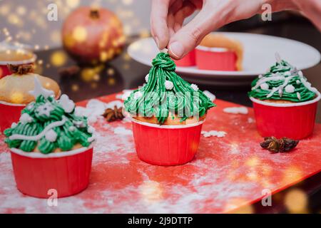 Christmas tree shaped cupcakes with cream frosting on christmas lights and festive background. Man hands Cooking Christmas cupcakes and decorate cupca Stock Photo