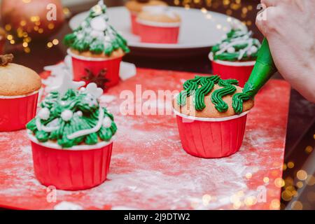 Christmas tree shaped cupcakes with cream frosting on christmas lights and festive background. Man hands Cooking Christmas cupcakes and decorate cupca Stock Photo