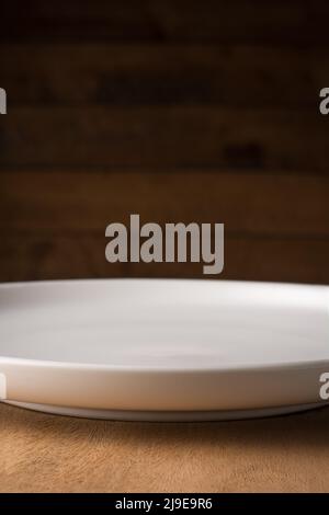 empty white plate on a wooden table top against dark blurry wooden background, food photography background or backdrop, closeup side view Stock Photo