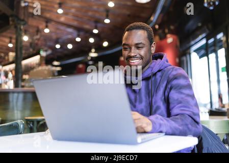 Positive African American male freelancer sitting at table and typing on netbook while working remotely in modern cafe Stock Photo