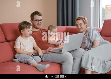 Big happy family watching movies playing games on laptop. Mother father kids spending time with gadgets on sofa Children with parents looking on Stock Photo