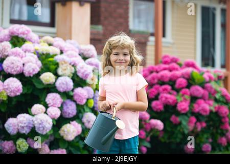 Cute little boy watering flowers with watering can in the garden. Child dressed in light summer closes and colourful t-shirt, smiling and having fun Stock Photo